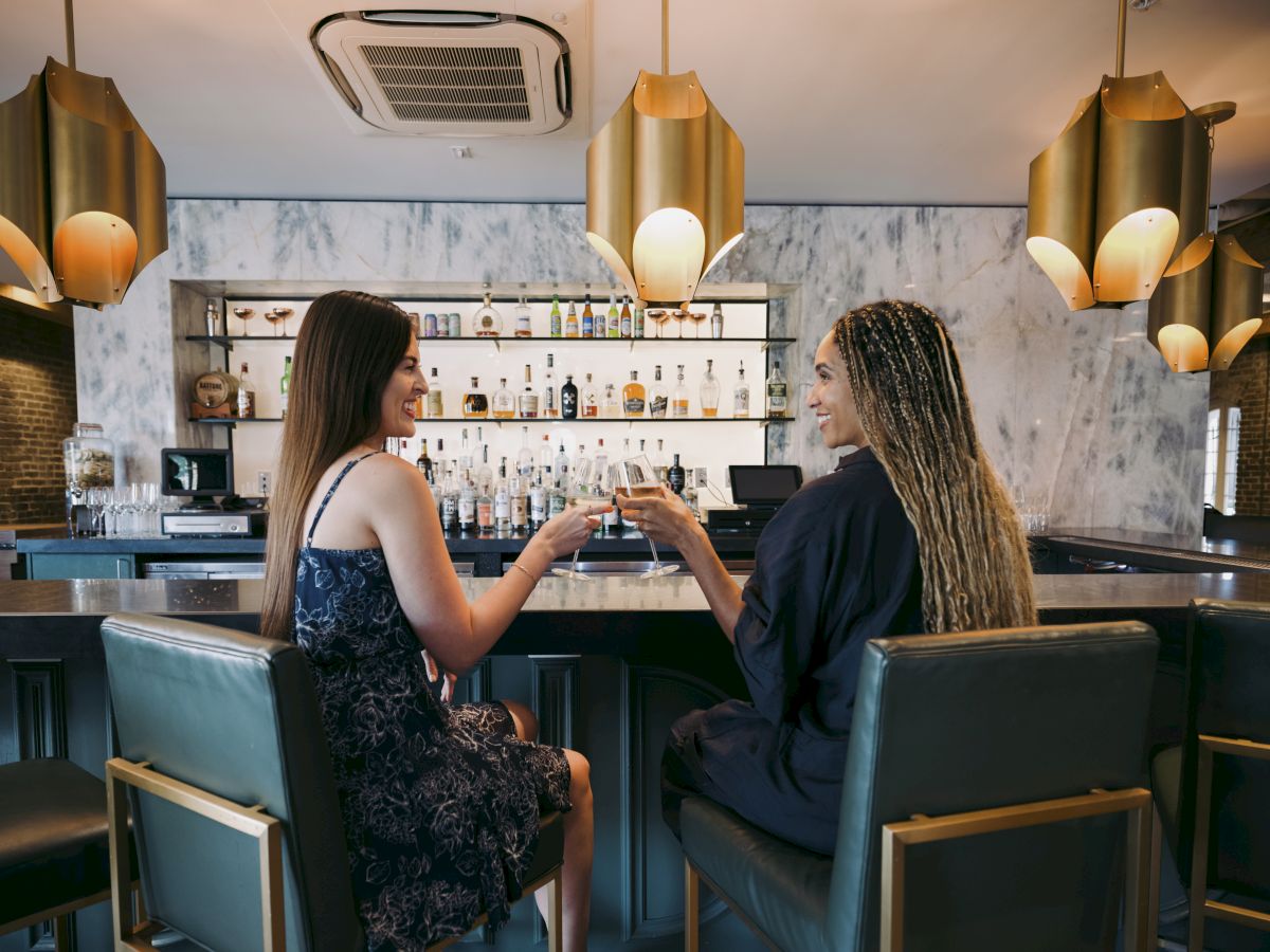 Two women are sitting at a bar, clinking glasses while smiling and talking. The setting is modern with gold decor and a well-lit background.