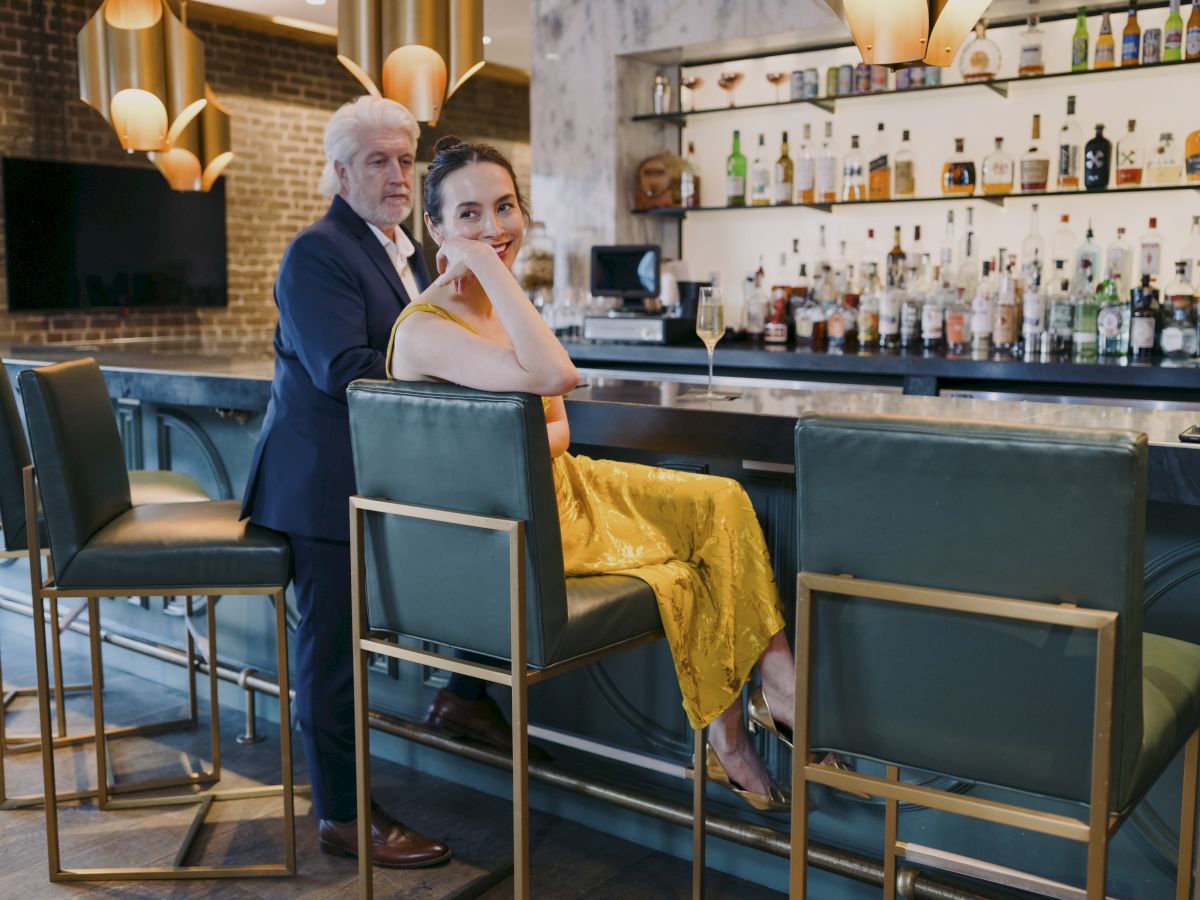 A man and a woman in formal attire sit at a stylish bar with modern decor, featuring shelves full of bottles and elegant lighting.