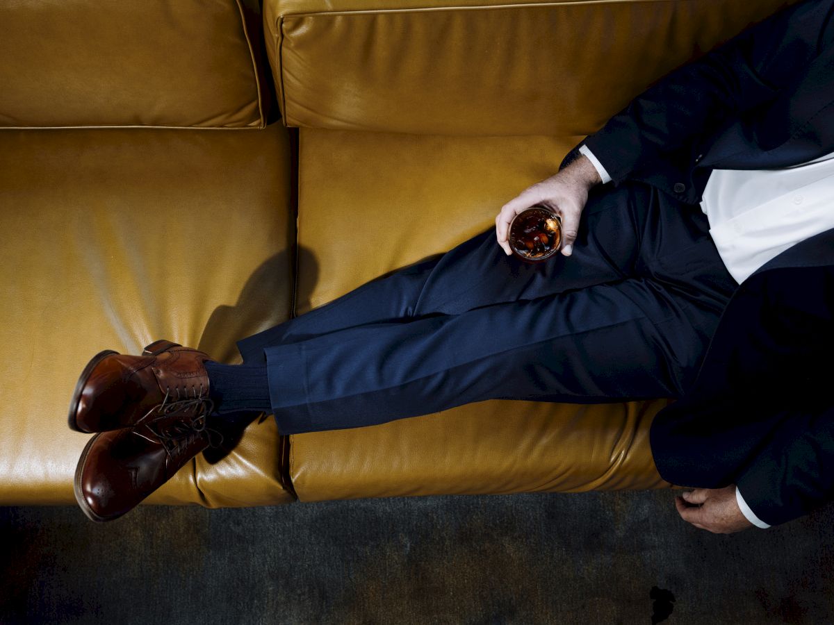 A man in a dark suit lounges on a tan leather sofa, holding a glass of liquor in his right hand with legs crossed over the seat's edge.