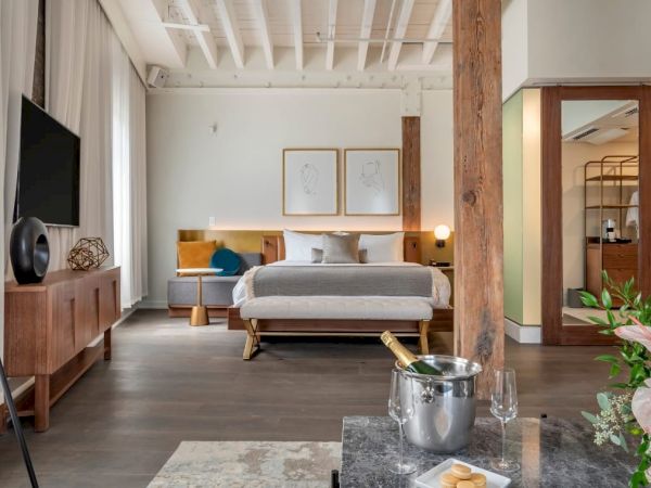 A modern bedroom with a bed, a TV, a sofa, artwork on the walls, a bathroom, flowers on a table, and champagne.