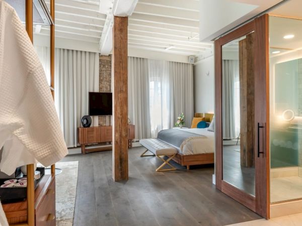 A modern hotel room features a bed, TV, glass shower, wooden beams, and a robe hanging near the entry.