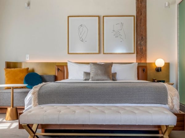 A modern bedroom features a neatly made bed with gray and white bedding, abstract wall art, and a cushioned bench at the foot of the bed.
