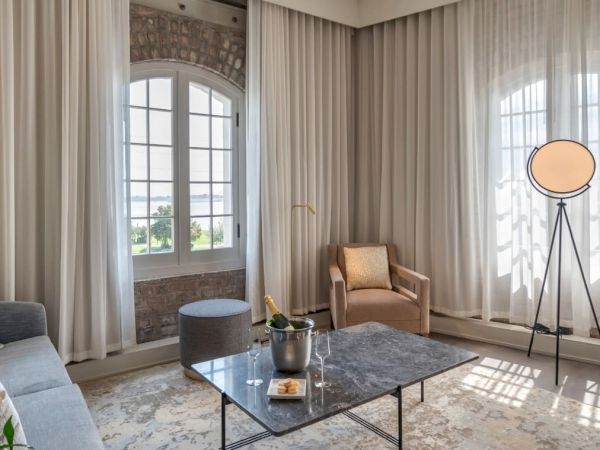 A modern living room with a sofa, armchair, coffee table with champagne bottle, tripod lamp, marble-top table, and tall windows with sheer curtains.