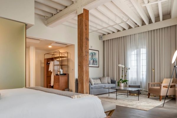 The image shows a modern, cozy hotel room with a bed, sitting area, wooden beams, and natural light through large windows.