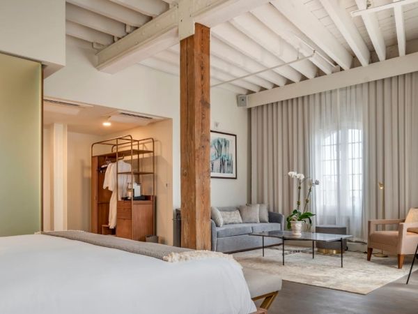 The image shows a modern, cozy hotel room with a bed, sitting area, wooden beams, and natural light through large windows.
