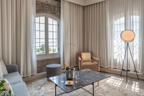 A modern living room with a sofa, armchair, coffee table with champagne bottle, tripod lamp, marble-top table, and tall windows with sheer curtains.