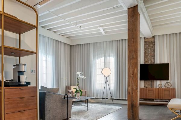 A modern living room with a cozy ambiance featuring a coffee maker, wooden furniture, a TV, a sofa, and a floor lamp next to large windows with sheer curtains.