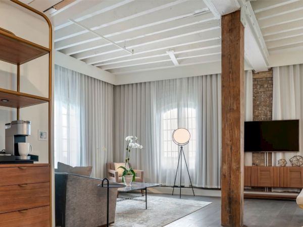A modern living room with a cozy ambiance featuring a coffee maker, wooden furniture, a TV, a sofa, and a floor lamp next to large windows with sheer curtains.