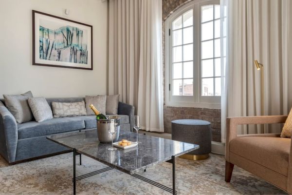 A cozy living room with a grey sofa, armchair, marble coffee table with champagne, an artwork, and a large window with curtains ending the sentence.