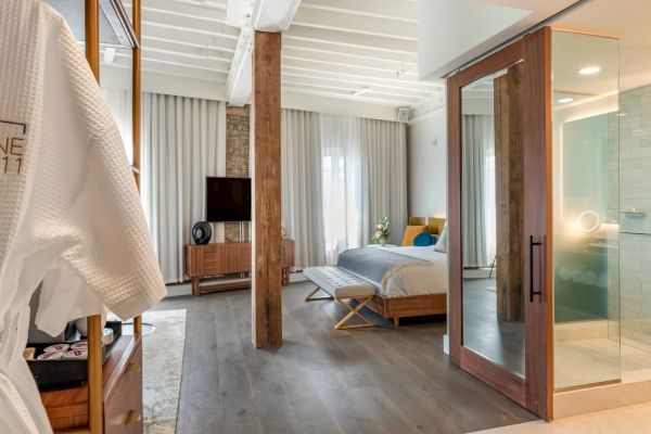 A modern hotel room features a bed, TV, glass shower, wooden beams, and a robe hanging near the entry.