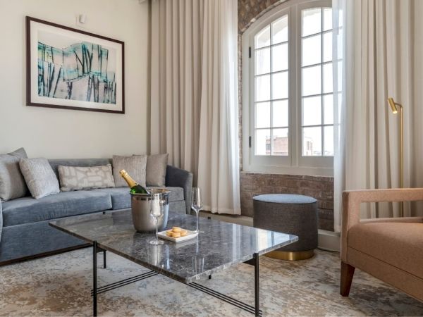 A cozy living room with a grey sofa, armchair, marble coffee table with champagne, an artwork, and a large window with curtains ending the sentence.