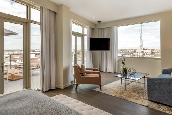 A modern living room with large windows, two armchairs, a sofa, a TV mounted on the wall, a coffee table, and a view of a balcony and cityscape.