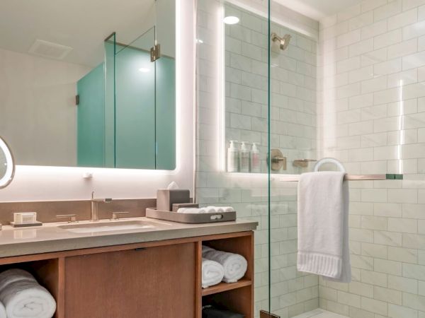 The image shows a modern bathroom with a vanity area, backlit mirror, a glass shower enclosure, white tiled walls, and neatly folded towels.