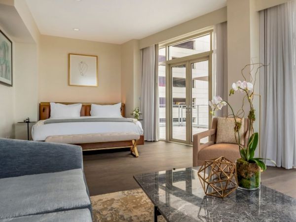 A modern hotel room with a bed, seating area, glass coffee table, and large windows overlooking a cityscape.