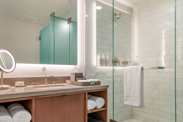 The image shows a modern bathroom with a vanity area, backlit mirror, a glass shower enclosure, white tiled walls, and neatly folded towels.