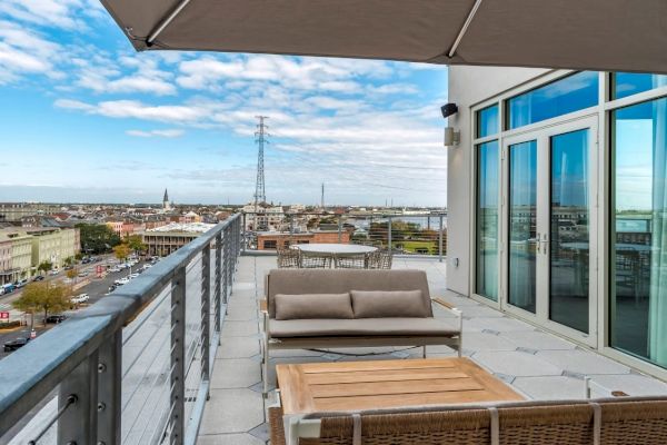 A rooftop terrace with modern outdoor furniture, overlooking a cityscape with buildings and a bridge under a partly cloudy sky.