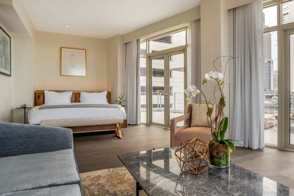 A modern hotel room with a bed, seating area, glass coffee table, and large windows overlooking a cityscape.