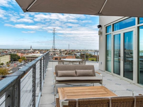 A rooftop terrace with modern outdoor furniture, overlooking a cityscape with buildings and a bridge under a partly cloudy sky.