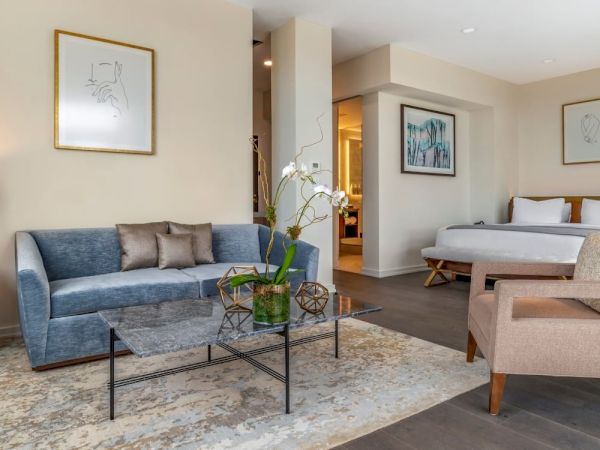 A modern hotel room features a seating area with a blue sofa, a coffee table, a bed, and a beige armchair, decorated with framed art.