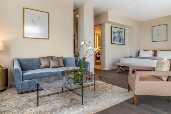 A modern hotel room features a seating area with a blue sofa, a coffee table, a bed, and a beige armchair, decorated with framed art.