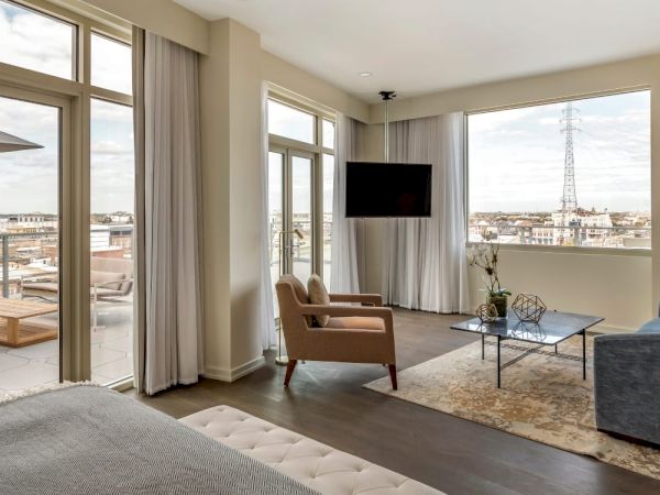 A modern living room with large windows, two armchairs, a sofa, a TV mounted on the wall, a coffee table, and a view of a balcony and cityscape.
