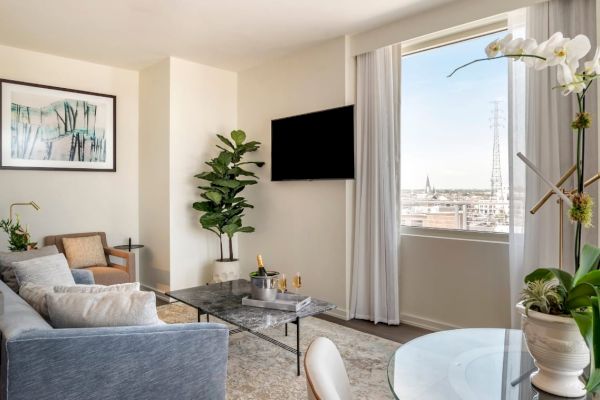 A modern living room with a couch, chairs, plants, a wall-mounted TV, artwork, a large window, and a glass coffee table with décor items.