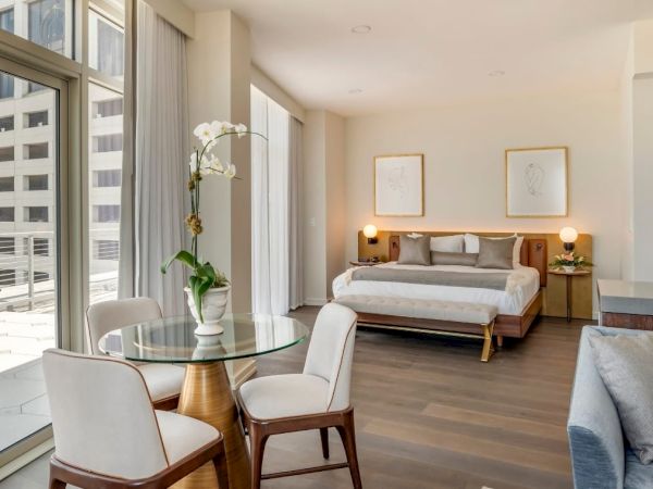A modern studio apartment features a bed, a seating area, a small dining table, and a balcony with city views. Warm lighting creates a cozy atmosphere.