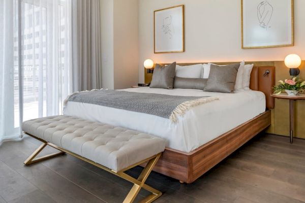 A modern bedroom with a large bed, two bedside tables, a cushioned bench, framed art, and a sheer curtain allowing natural light.