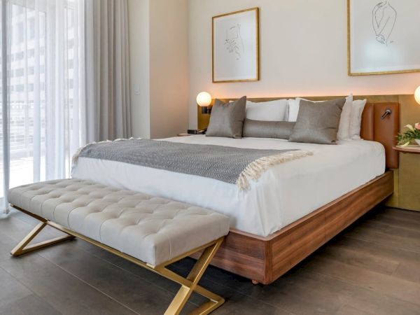 A modern bedroom with a large bed, two bedside tables, a cushioned bench, framed art, and a sheer curtain allowing natural light.