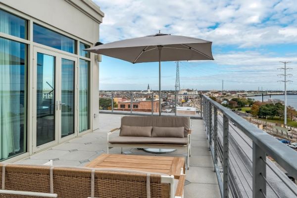 An outdoor patio with a sofa, chairs, table, and umbrella overlooks a cityscape and a body of water. The sky is partly cloudy.