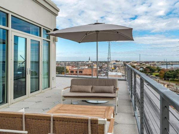 An outdoor patio with a sofa, chairs, table, and umbrella overlooks a cityscape and a body of water. The sky is partly cloudy.