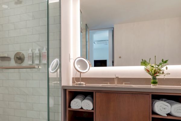 A modern bathroom with a glass shower, double sinks, rolled towels, a vanity mirror, and a small vase with flowers on the counter.