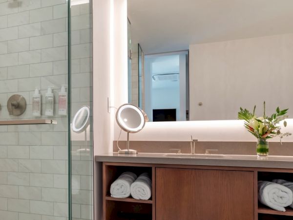 A modern bathroom with a glass shower, double sinks, rolled towels, a vanity mirror, and a small vase with flowers on the counter.