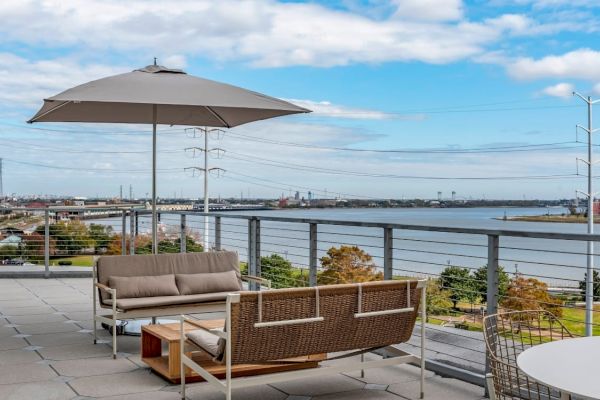 The image shows a rooftop patio with seating, an umbrella, and a scenic view of a river and cityscape under a partly cloudy sky.