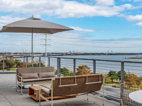 The image shows a rooftop patio with seating, an umbrella, and a scenic view of a river and cityscape under a partly cloudy sky.