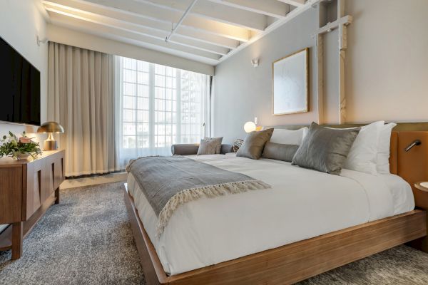 A modern, cozy bedroom with a large bed, wooden furniture, a seating area near a large window, and soft lighting, creating a relaxing ambiance.