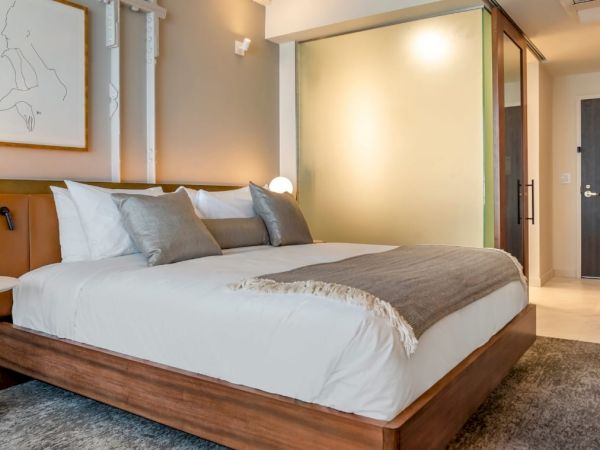 A modern hotel room features a neatly made bed with pillows, wall art, bedside lamps, and a frosted glass partition leading to the bathroom.