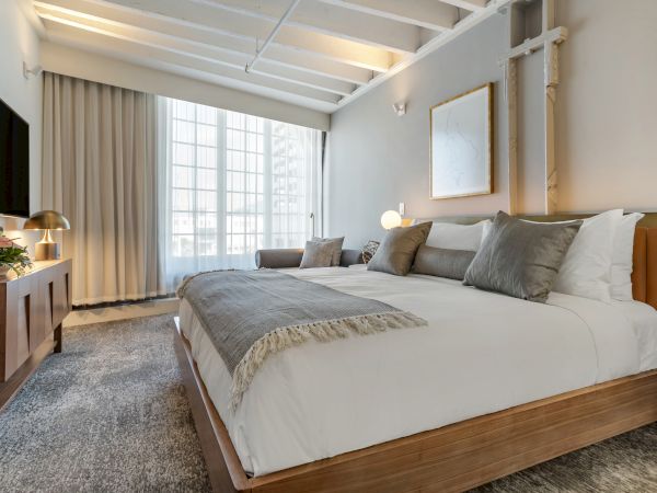 A modern, cozy bedroom with a large bed, wooden furniture, a seating area near a large window, and soft lighting, creating a relaxing ambiance.
