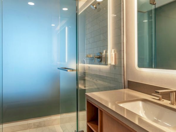 The image shows a modern bathroom with a glass shower enclosure, a white sink and countertop, and stylish lighting on a mirror.
