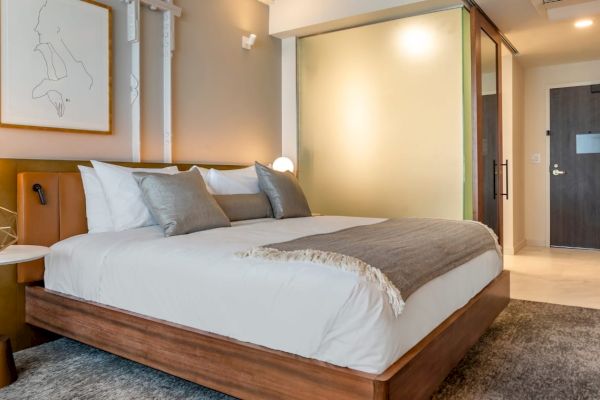 A modern hotel room features a neatly made bed with pillows, wall art, bedside lamps, and a frosted glass partition leading to the bathroom.