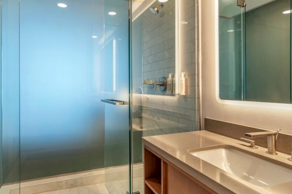 The image shows a modern bathroom with a glass shower enclosure, a white sink and countertop, and stylish lighting on a mirror.