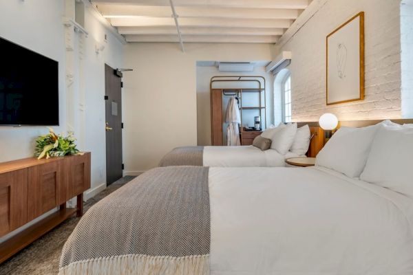 A modern hotel room with two neatly made beds, a wall-mounted TV, a wooden dresser, and warm lighting situated in a bright, minimalist design ending the sentence.