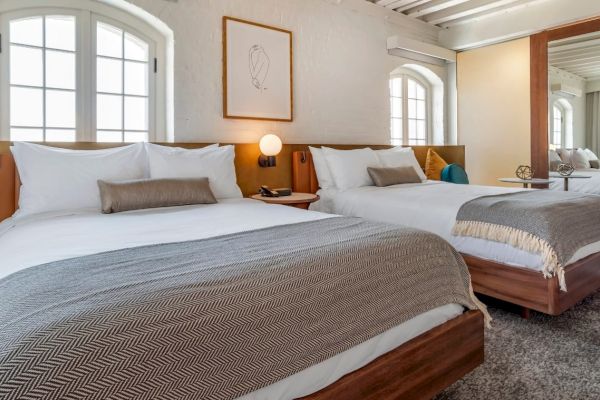 The image shows a cozy bedroom with two neatly made beds, modern decor, a mirrored closet, and two large windows letting in natural light.