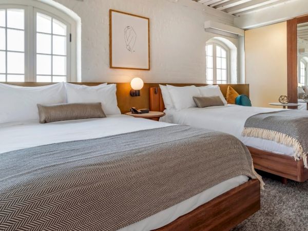 The image shows a cozy bedroom with two neatly made beds, modern decor, a mirrored closet, and two large windows letting in natural light.