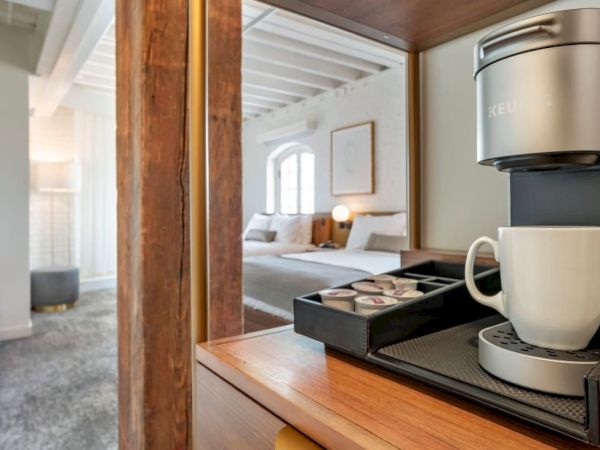 A modern hotel room with a coffee maker on a wooden shelf, a mug, assorted coffee pods, and a neatly made bed in the background.