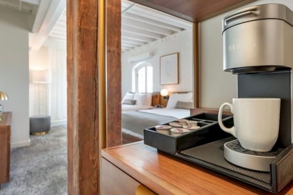 A modern hotel room with a coffee maker on a wooden shelf, a mug, assorted coffee pods, and a neatly made bed in the background.