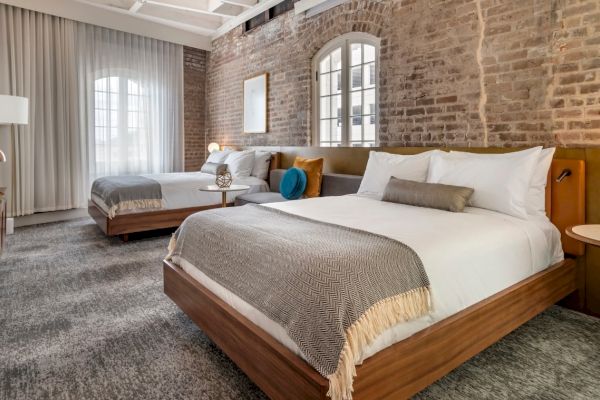 The image shows a stylish hotel room with two large beds, a brick wall, modern light fixtures, and a window with sheer curtains.