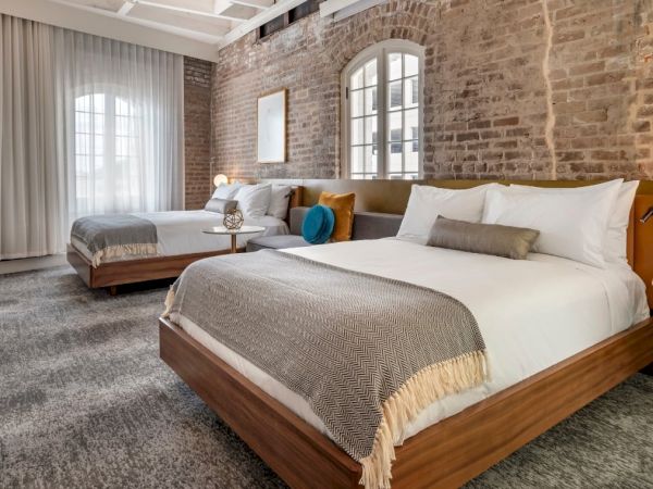 The image shows a stylish hotel room with two large beds, a brick wall, modern light fixtures, and a window with sheer curtains.