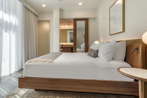 Modern bedroom with a wooden bed, white linens, bedside table, mirror closet, white walls, and a large window with curtains.