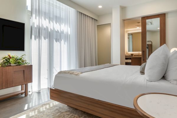 A modern bedroom featuring a large bed with white linens, a wooden nightstand with a plant, and a TV mounted on the wall.